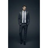 En mand iført gråt jakkesæt, hvid skjorte og sort slips står med hænderne i lommerne mod en mørk baggrund.