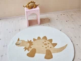 På et lyst trægulv ligger et rundt hvidt tæppe med et tegneseriemotiv af en stegosaurus. I nærheden står en lyserød skammel med et stegosaurus-plysdyr.