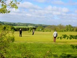 Tre personer spiller golf på en grøn mark under en delvis overskyet himmel med træer og bakker i baggrunden. En person gør klar til at putte, mens de andre ser på.
