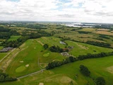 Luftfoto af en golfbane omgivet af grønne marker, træer, en parkeringsplads, bygninger, en dam og fjerne bølgende bakker under en delvist overskyet himmel.