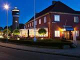 Et murstenshotel med en oplyst glasindgang og udeservering ses i skumringen ved siden af et højt cylindrisk tårn og sirligt klippede buske.