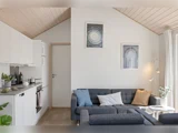 Moderne stue med grå modulsofa, pastelfarvede puder, et lille rundt bord med blomster, hvide vægge, indrammede kunstværker og et køkkenområde til venstre. Loft og gulv i lyst træ.