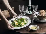 En person serverer et fad med salat og kogte æg ved et dækket spisebord med glas, brød i en kurv og en lille skål med dressing.
