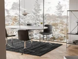Moderne spiseplads med et rundt hvidt bord, fire sorte stole og et sort tæppe ved et stort vindue med udsigt til sneklædte træer og bjerge. Der står et glas vin på bordet.