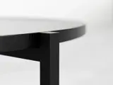 Nærbillede af kanten og benet på et moderne sort metal- og træbord på en almindelig, lys baggrund.