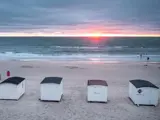 Fire små hvide strandhytter på sand ligger ud til et roligt hav ved solnedgang, med nogle få mennesker nær vandet og en overskyet himmel over.