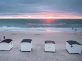 Fire små hvide strandhytter på sandet vender ud mod havet ved solnedgang, mens bølgerne ruller ind og himlen er overskyet.