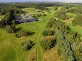 Luftfoto af en grøn golfbane omgivet af træer, med bygninger og stier i midten og skovområder i baggrunden.