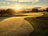 Solopgang over en golfbane, der kaster varmt lys og lange skygger over det duggede græs, sandbunkerne og flagstangen i det fjerne.