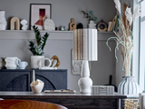 Et moderne skrivebord med en hvid lampe, lysestage, kuglepen og stribet æske i et rum med dekorative genstande, planter og indrammet kunst på hylder i baggrunden.