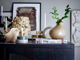 En moderne hylde viser abstrakte vaser, stearinlys, bøger og bladgrønt i en vase med indrammet kunst og tekstureret indretning i baggrunden.