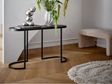 En moderne entré har et sort konsolbord med bøger, en abstrakt sølvskulptur og en vase med grønne planter ved siden af en struktureret bænk og et rundt tæppe på et sildebensgulv.
