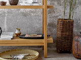 På træhylder står keramik, bøger og en bakke ved siden af en høj flettet kurv med planter, der står op ad en struktureret betonvæg. En dekorativ rund kurv står på gulvet.