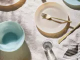 Et bord med pastelfarvede tallerkener og skåle, en guldgaffel og -kniv, et klart glas vand og en stribet stofserviet på en hvid linneddug.