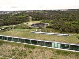 Luftfoto af en moderne bygning med græstag og store glasvinduer ved siden af en cirkulær betonkonstruktion omgivet af træer og grønne områder.