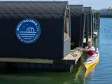 En mand i en gul kajak nærmer sig moderne flydende hytter med "Boat Houses DK"-logo på siden på et roligt vand.