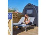 En kvinde sidder i en liggestol på en træterrasse og klapper en lille hund i nærheden af en lille moderne hytte og et skilt med teksten Boathouse NR.3.