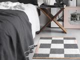 Et sort- og hvidternet tæppe ligger på gulvet ved siden af en seng med et mørkt tæppe; et træstativ og indrammede kunstværker står i nærheden.