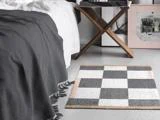 Et ternet tæppe med grå, hvide og beige firkanter ligger på gulvet ved siden af en seng med et mørkegråt tæppe med frynser og en træskammel med en bakke.