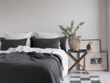 Et moderne, minimalistisk soveværelse med en pænt redt seng med hvidt og mørkegråt sengetøj, et lille sidebord i træ med en potteplante og indrammet kunst, der hviler på gulvet.
