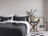 En pænt redt seng med hvide og mørkegrå puder, et gråt tæppe, et lille sidebord i træ med en potteplante og indrammet kunst, der læner sig op ad en hvid væg.