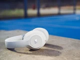 Hvide hovedtelefoner hviler på en betonoverflade med en blå udendørs sportsbane og et kædehegn sløret i baggrunden.