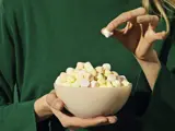 En person i en grøn sweater holder en skål med farverige mini marshmallows og tager en op med fingrene.