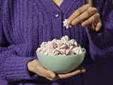 En person i en lilla sweater holder en skål med lyserøde marengskager og tager en op med højre hånd.