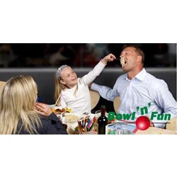 En pige fodrer legende en mand med pasta, mens en kvinde ser på og smiler. De sidder ved et bord med mad på et sted, der hedder Bowl n Fun.