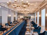 Elegant restaurantinteriør med blå fløjlsstole, lange banketter, dækkede borde, moderne lysekroner og en velassorteret bar bagved.