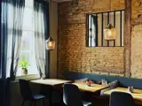 Hyggelig café med murstensvægge, træborde, sorte stole, hængende pendler, et vindue med gennemsigtige gardiner og en lille plante i vindueskarmen.