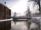 En murstensbygning ved en rolig flod, en træbro og bladløse træer, alt sammen dækket af et tyndt lag sne under en overskyet vinterhimmel.