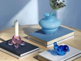 Et rundt træbord med stablede bøger, en blå vase med lavendel, et hvidt lys i en lyserød holder og en lille blå glasskulptur, alt sammen oplyst af blødt sollys.