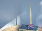 Et højt, tyndt stearinlys i en lyserød glasholder står på en lukket blå bog, som er stablet på en anden bog på et lyst træbord mod en lyseblå væg.