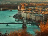 En naturskøn udsigt over Budapest med Donau-floden, parlamentsbygningen, broer, både og efterårsfarvede træer i forgrunden.