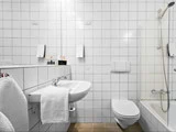 Et hvidt flisebadeværelse med håndvask, væghængt toilet, håndklæde, spejl, toiletartikler og et badekar med glasbruser og brusehoved.