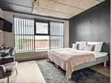 Et moderne hotelværelse med en stor seng, grå vægge, stort vindue med lodrette persienner, væghængt tv, skrivebord med stol og et blødt tæppe på gulvet.