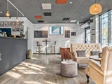 Moderne caféinteriør med hyggelige sofaer, lænestole, et lille rundt bord, vægkunst og store vinduer, der lukker naturligt lys ind. En disk er synlig til venstre.