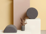 To grå stofbetrukne geometriske puder, en rund og en halvcylindret, er placeret på hvide blokke ved siden af en lille grå vase med tørret græs på en beige og ferskenfarvet baggrund.