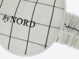 Nærbillede af en øjenmaske af hvidt stof med et sort gittermønster og ordene by NORD broderet på.