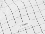 Nærbillede af et hvidt stof med et sort gittermønster og en hvid label, hvor der står by NORD, syet på.