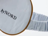 En øjenmaske i lyseblåt stof med brun kant og teksten byNORD broderet på, med en rynket elastikstrop.