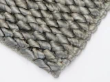 Nærbillede af et hjørne af et vævet, gråt tekstureret tæppe eller måtte med tykke, flettede fibre, placeret på en hvid overflade.