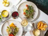 To tallerkener med ristede grøntsager og mikrogrønt, glas med rød- og hvidvin, pommes frites, sauce i en lille gryde og vandglas på en hvid dug.