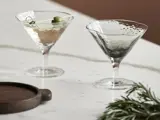 To strukturerede martiniglas på en marmorflade; det ene indeholder en lys drink med en oliven, det andet er tomt. En træbakke og rosmarinkviste står i nærheden.