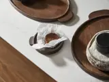 En rund chokoladekage ligger på krøllet hvidt pergamentpapir oven på en lille træbakke, omgivet af større serveringsbrætter af træ på en hvid bordplade.