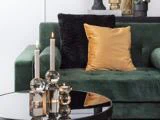 En grøn fløjlssofa med en guldpude og en sort pude bag et sofabord af glas med tændte stearinlys og et fyrfadslys.
