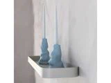 To hvide bloklys i blå, skulpturelle lysestager står på en minimalistisk, rektangulær hvid væghylde mod en lys væg.