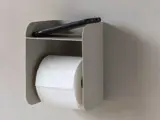 En grå væghængt toiletpapirholder med en rulle toiletpapir og en smartphone på den øverste hylde.