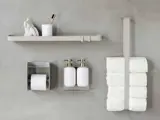 Minimalistisk badeværelsesvæg med en hylde, sæbedispenser, tandbørsteholder, toiletpapirholder og stablede toiletruller i en metalholder mod en grå baggrund.